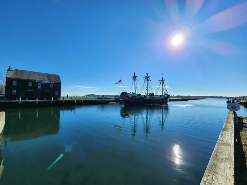 A historic ship is seen on the water with a bright sun overhead and a deep blue sky