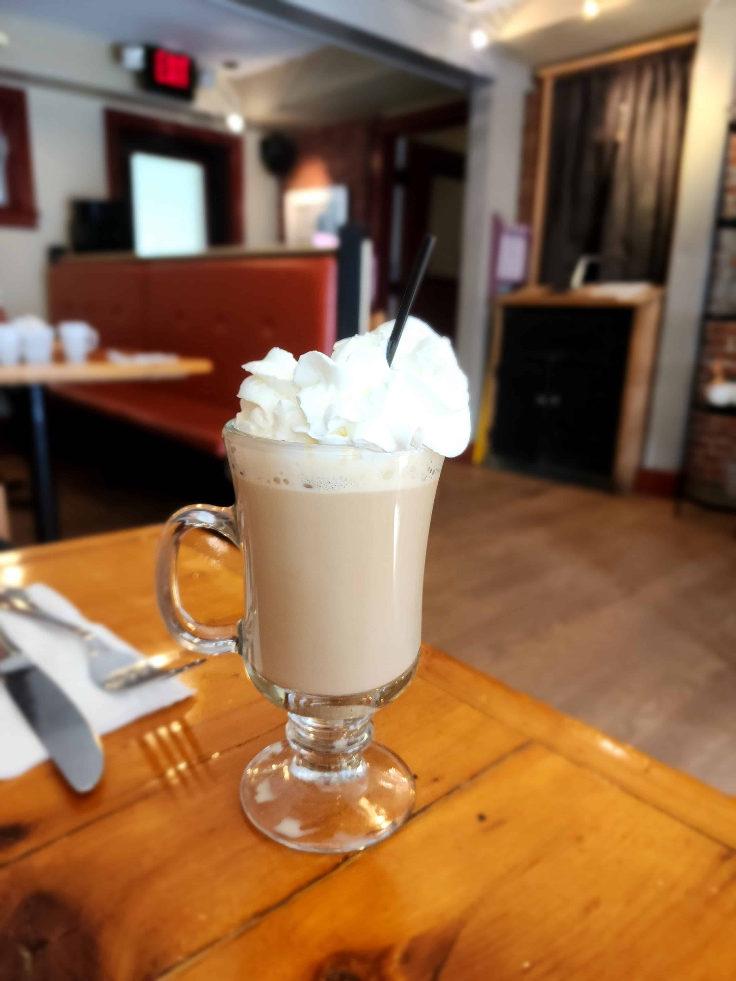 a hot Irish coffee topped with whipped cream sits in a glass mug on top of a restaurant table, background is blurred