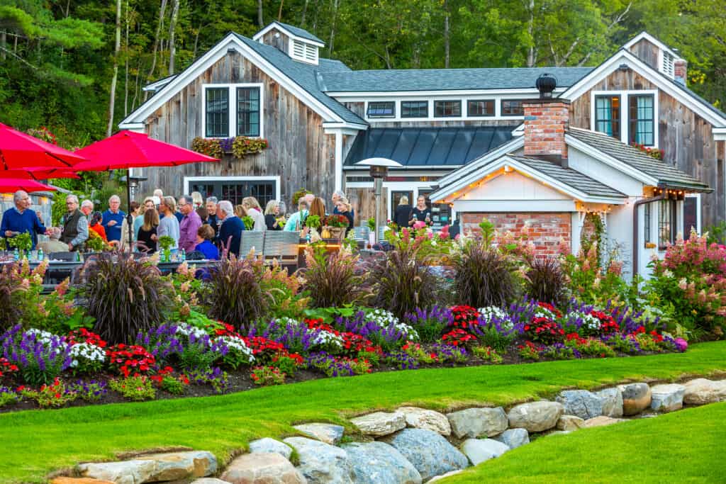 A well landscaped lawn with colorful flowers and a rock trench sit in front o fa large inn with many people attending a party.