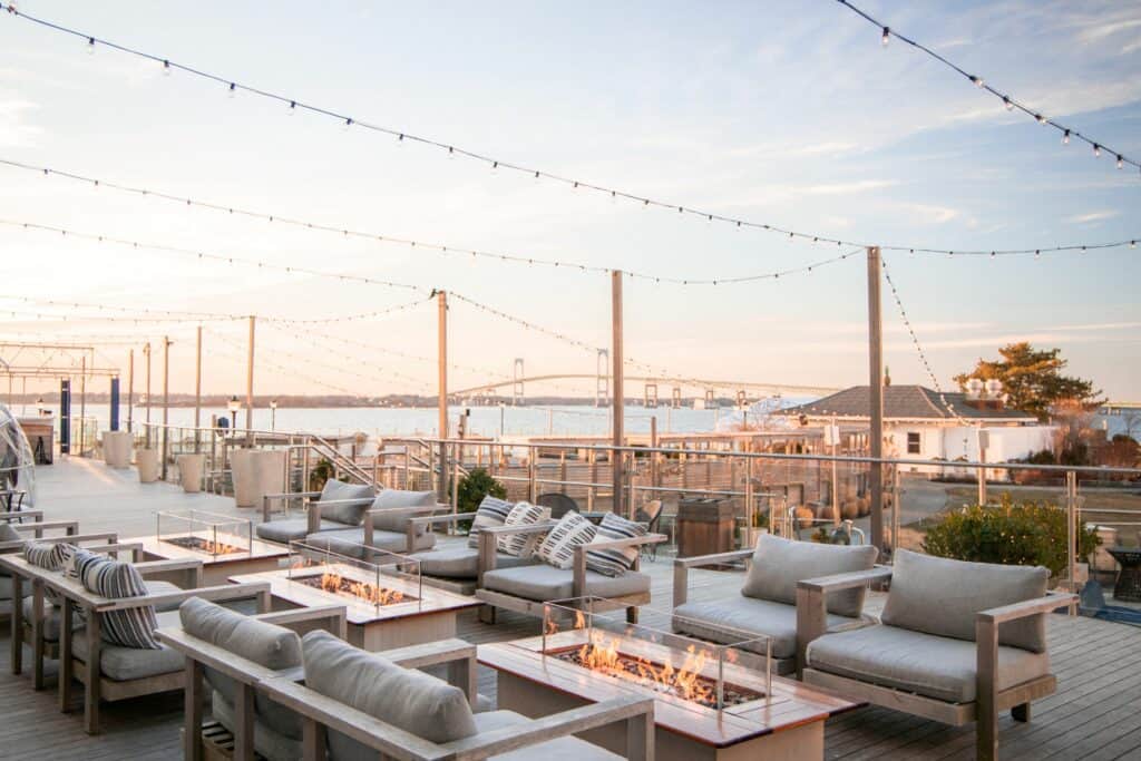 An outdoor fire pit space at Newport Harbor Island Resort is seen with seating, overhead lighting, and the iconic Newport Bridge in the background.