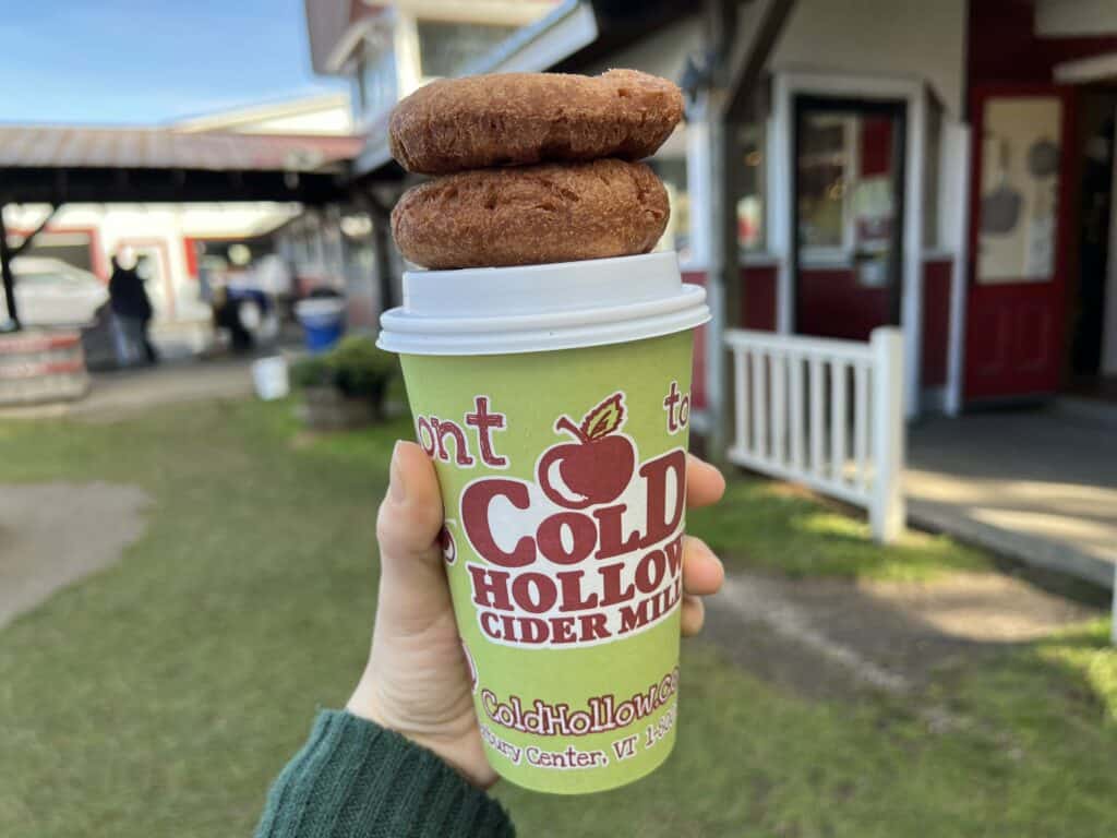 hand holding a lime green coffee cup that says cold hollow cider mill on it. cup is topped with two cider donuts