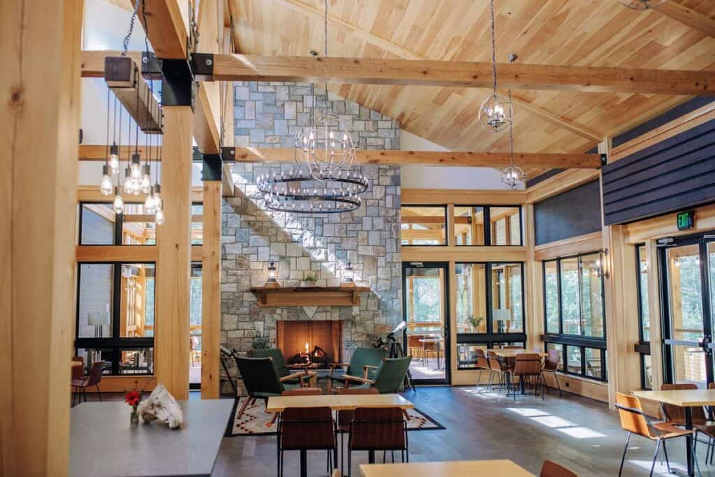 A large lodge is seen with windows surrounding and natural light streaming in on tables and a floor to ceiling fireplace
