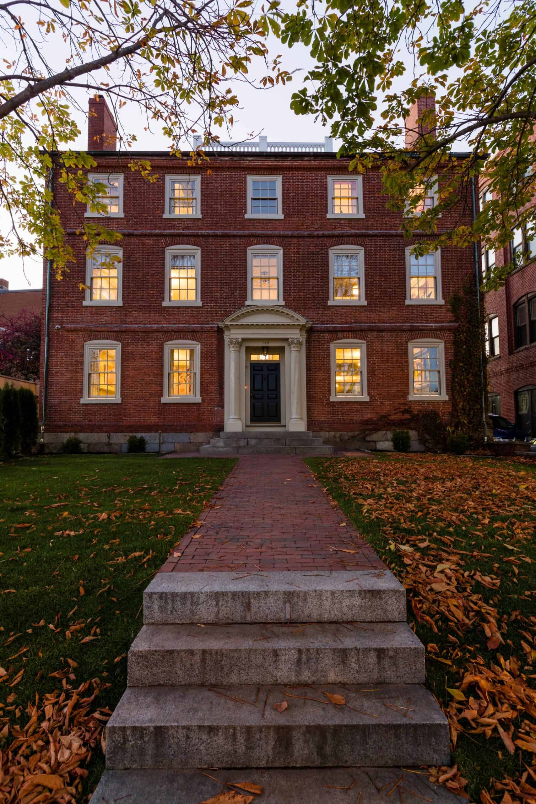 A somewhat imposing historical haunted hotel made of red bricks is seen on a fall day with yellow-brown leaves scattered over the lawn.