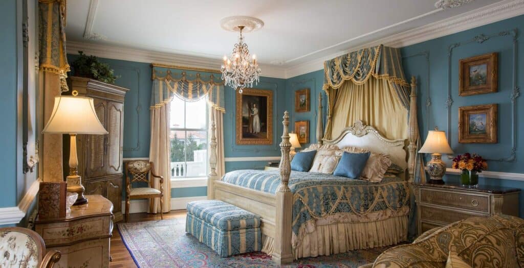 An intricately decorated elegant suite at one of the top luxury Newport RI hotels is seen with blue bedding and walls and a chandelier hanging overhead