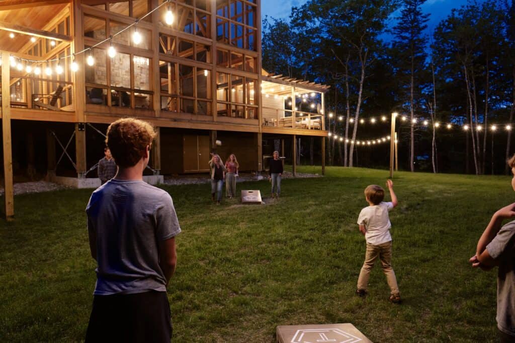 A group of people enjoys the yard in front of an outdoor resort in Acadia Nationl Park at dusk with trees and lights surrounding the yard.