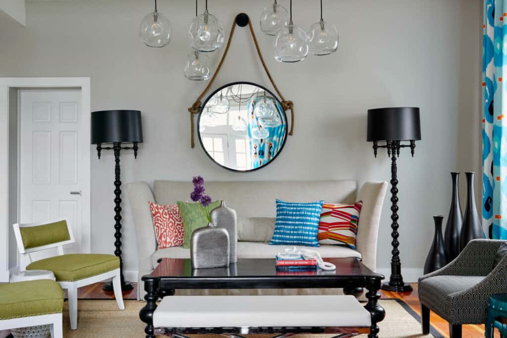 A couch is seen with two matching black lamps on either side, and colorful funky pillows with a decorative mirror and chandelier above