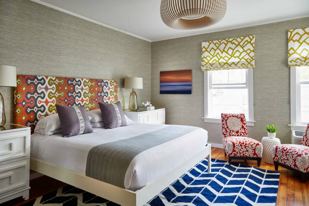 A queen sized bed with a colorful headboard, rug beneath, and side chair sit beside a window in one of the top boutique hotels in Newport, Rhode Island