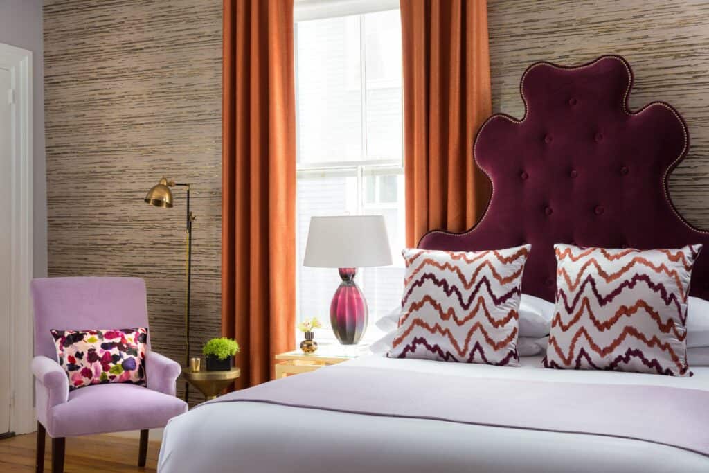 A guest room at The Gilded in Newport, Rhode Island is seen with a deep fuscia headboard, purple chair and matching side lamp