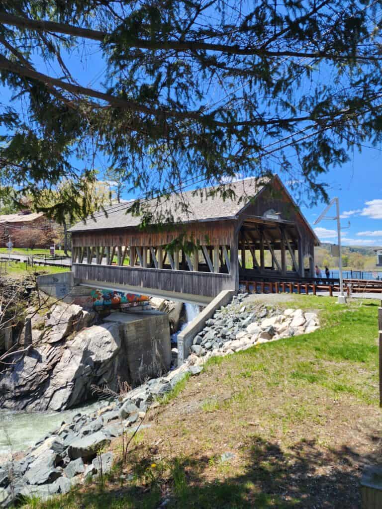 a brown wooden covered bridge sits over a rock sided river. a pine tree's branches border the image in the foreground