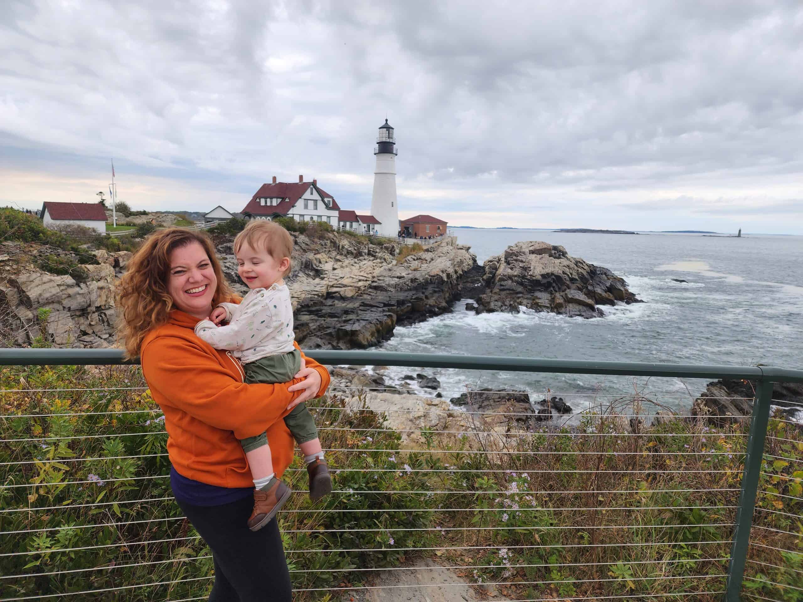 amy stands in front of an iconic lighthouse, portland maine, holding a toddler. both are smiling