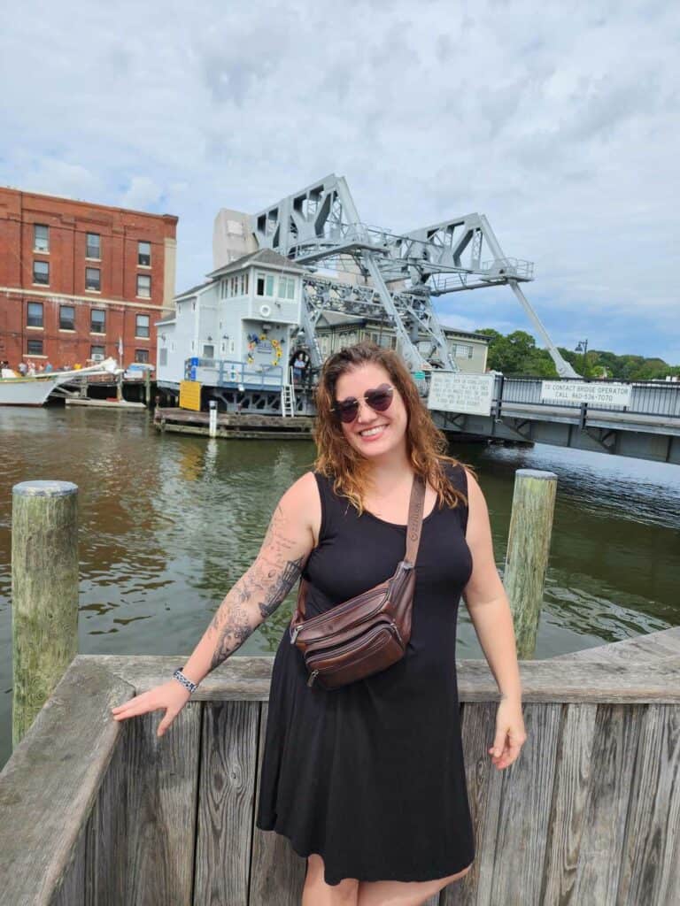 Amy, a brown haired 30 something woman stands in a black dress in front of a drawbridge 