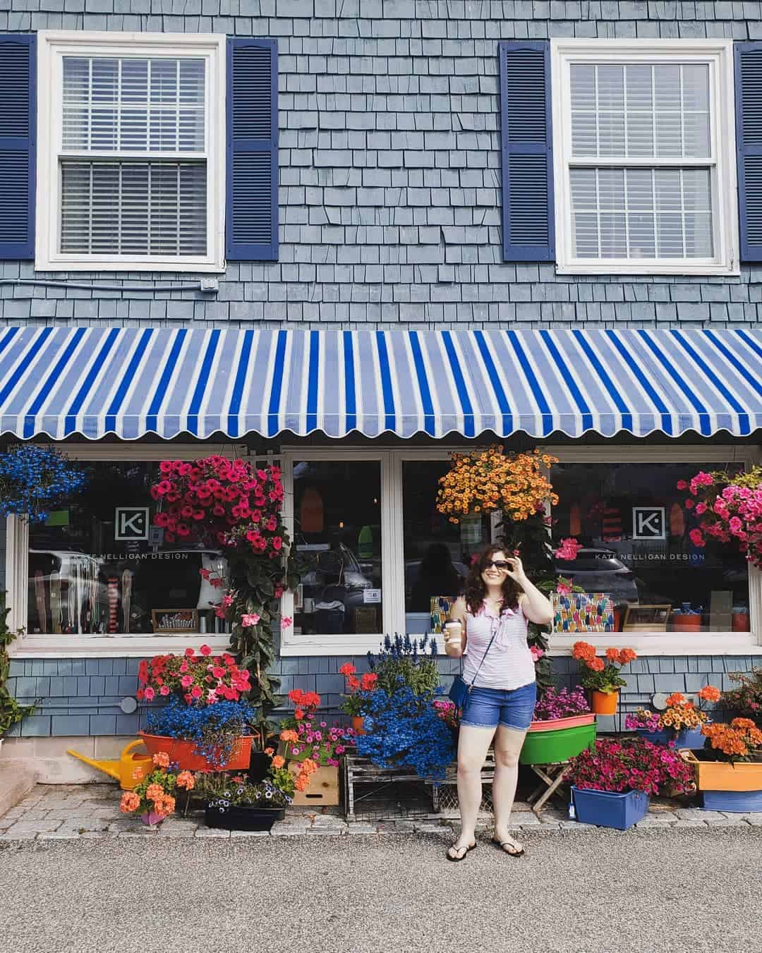 amy standing in front of a shop with bright flowers, she wears shorts, a tank top and sunglasses