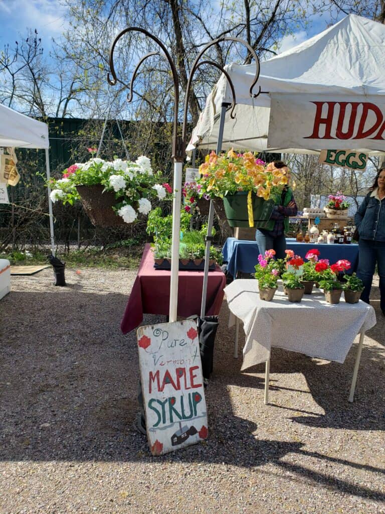 a table at a farmers market showing fresh flowers. a hand painted sign for pure maple syrup on the ground in front