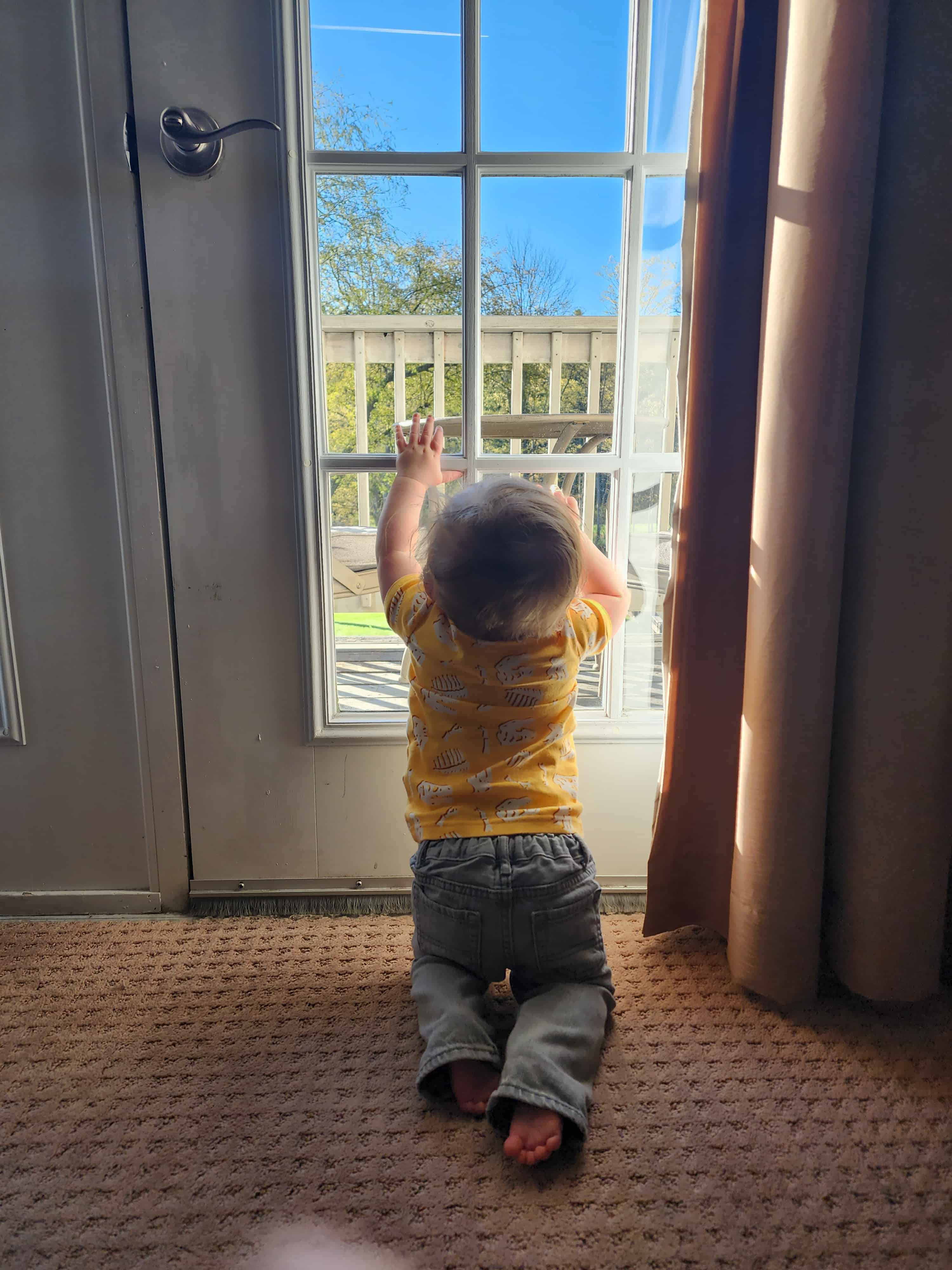 a young toddler kneels against a french balcony door looking out onto a blue sky