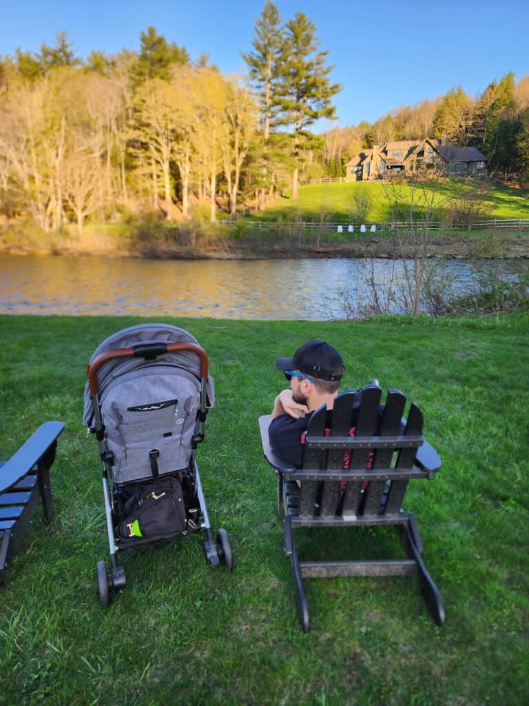 a man sits in an adirondack chair facing a river while a baby stroller sits next to him pointing in the same direction