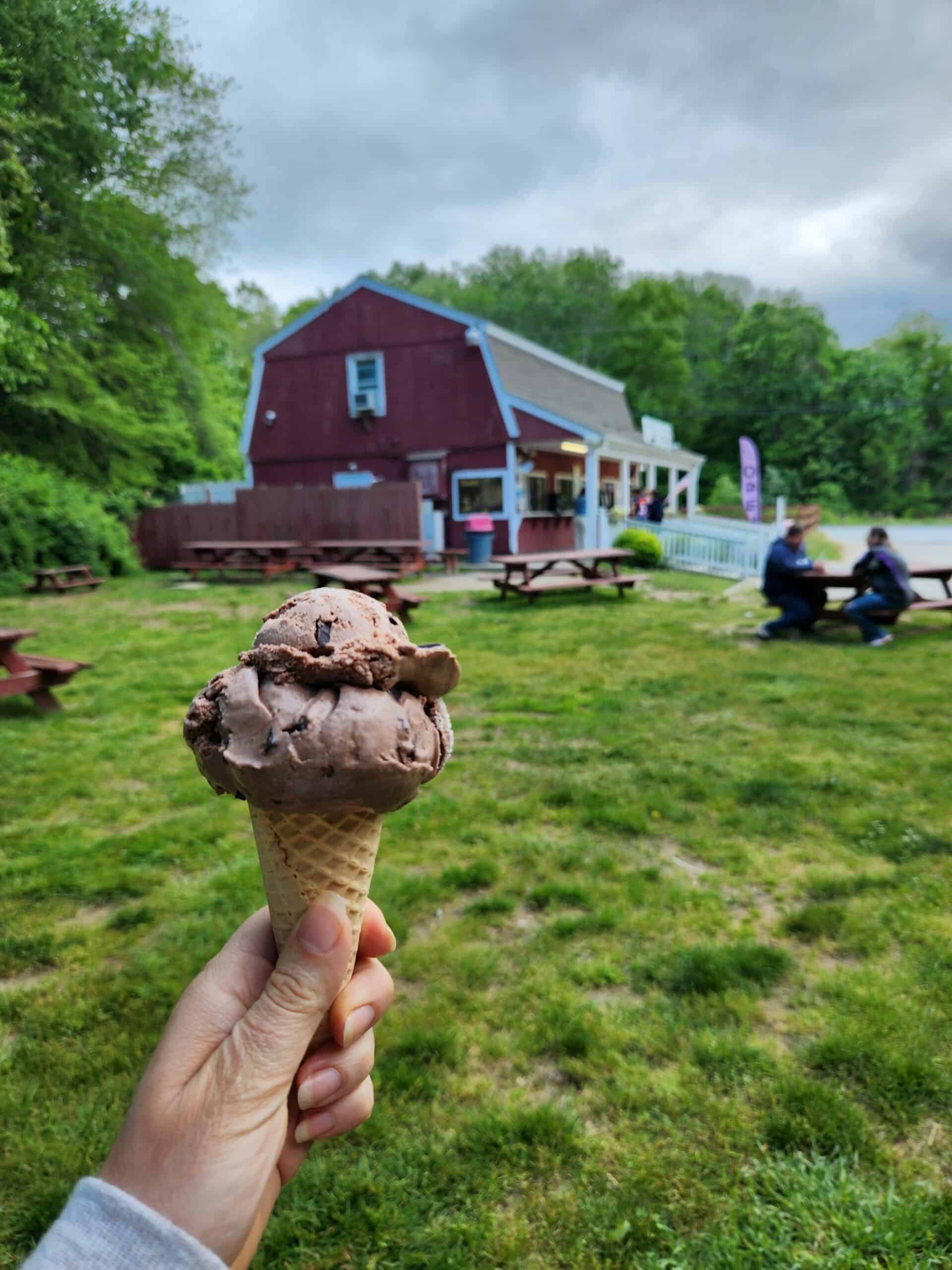 An outstretched hand holding a chocolate ice cream cone with a large red barn in the background 