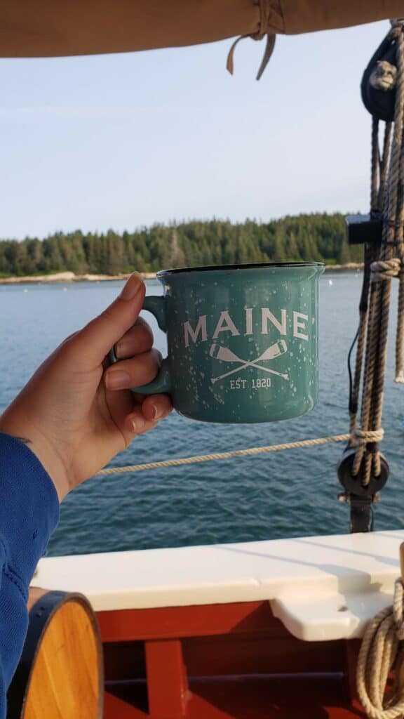 A hand holding a mug that says Maine against the backdrop of a heavily forested shore