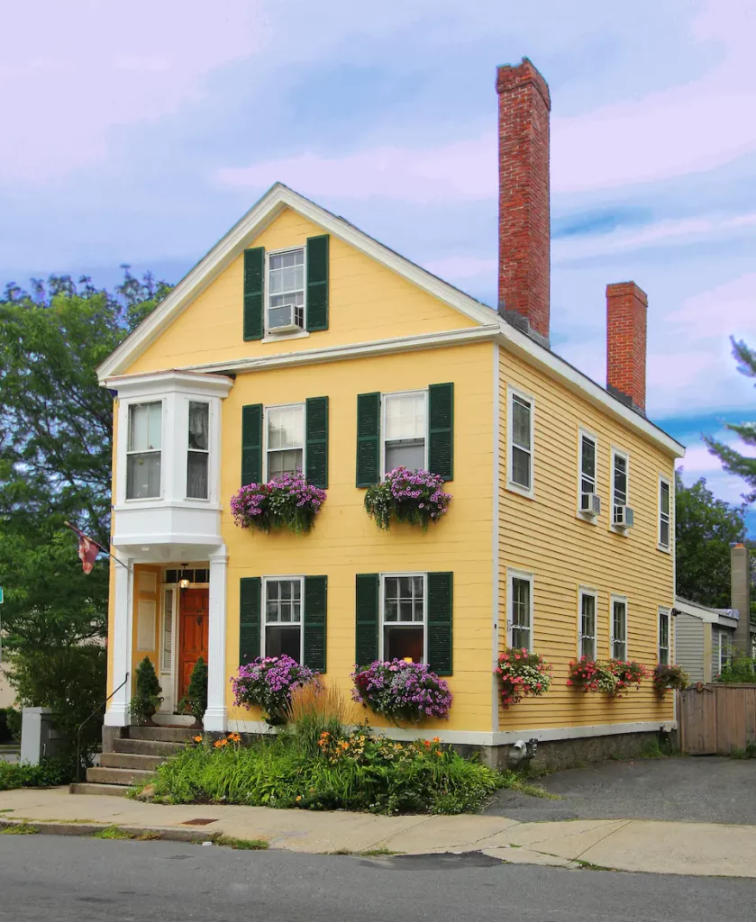 exterior of an 1800s house painted yellow with green shutters and flowers in each window box