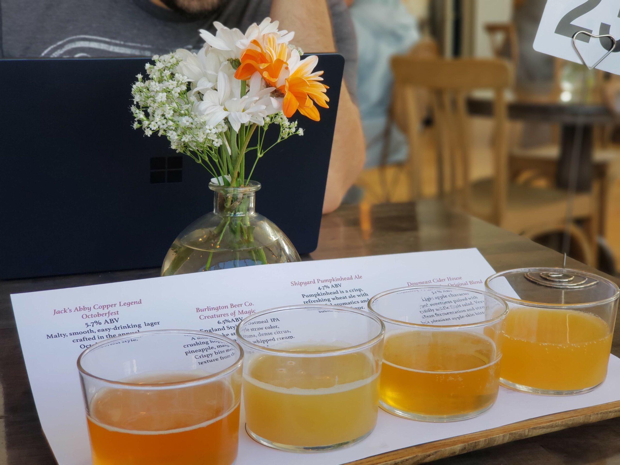 This image depicts a flight of five craft beers, each in a small glass, arranged on a table with a menu or beer description sheet underneath. In the background, there is a small vase with a bouquet of white daisies and orange flowers. A person is sitting behind a laptop, partially visible, creating a casual atmosphere in what appears to be a brewery or café setting