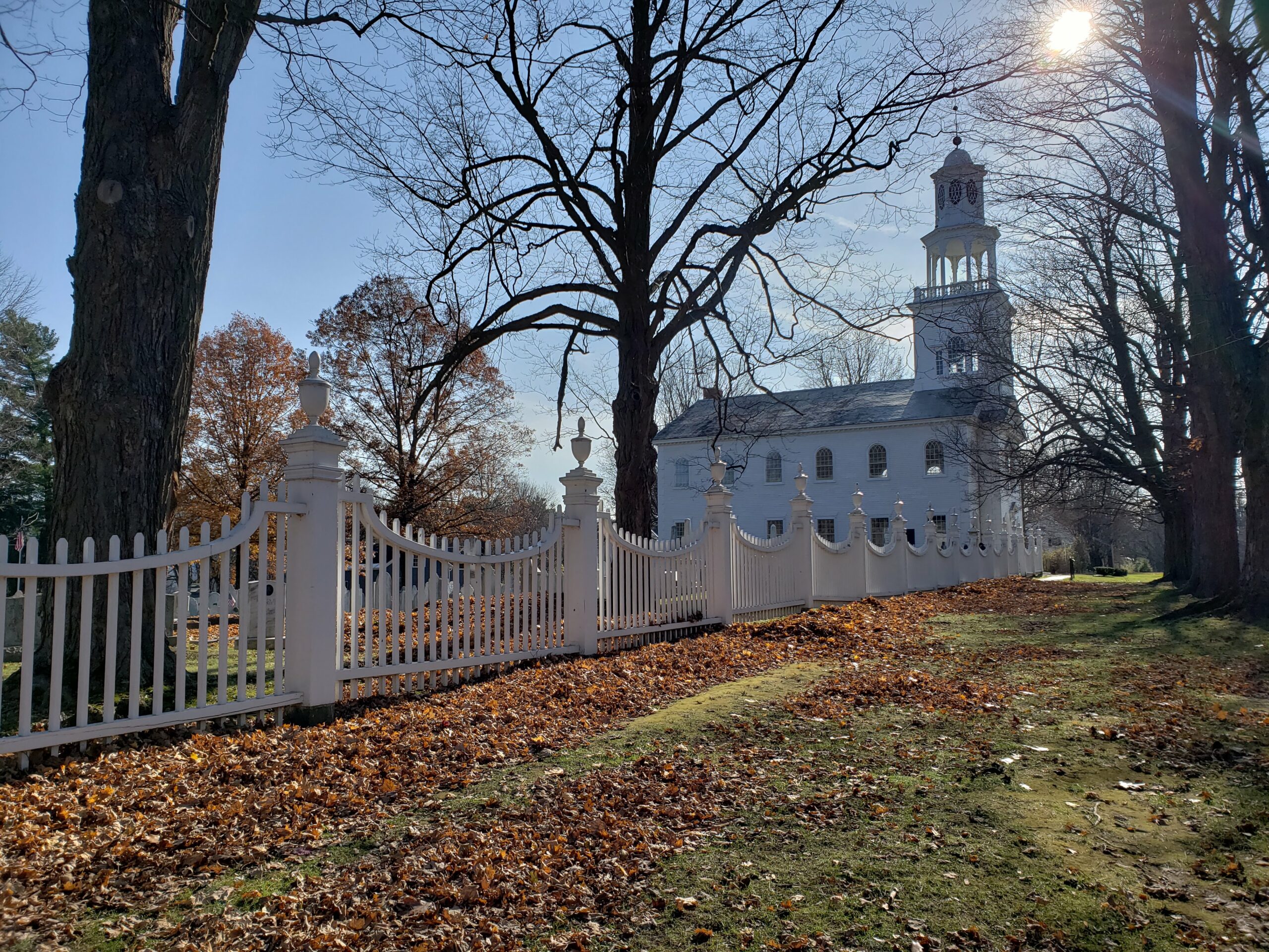 This image shows a bright, sunny day with a historical white church featuring a clock tower in the background. The church is surrounded by a decorative white picket fence with ornate posts, and the ground is covered with fallen autumn leaves. Bare trees line the fence, casting shadows across the path as sunlight filters through the branches.