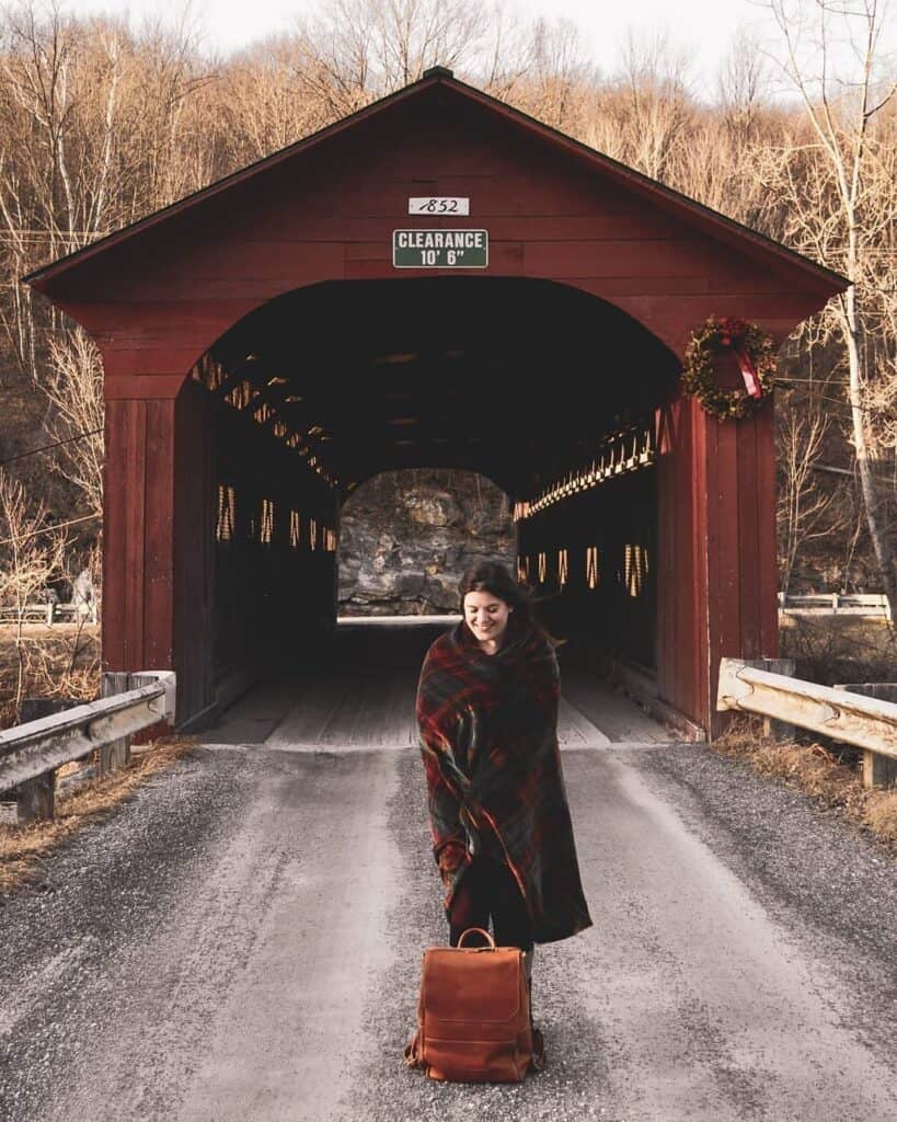 amy wrapped in a red blanket stands in front of a red covered bridge with a christmas wreath on it