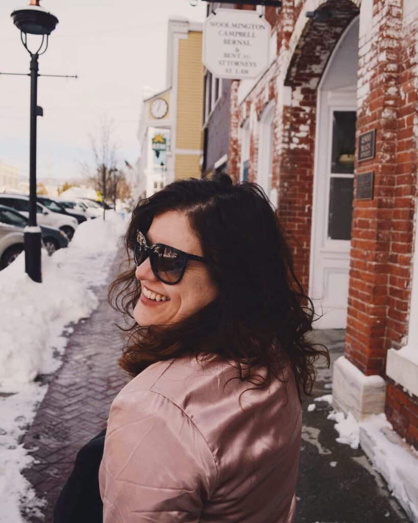 amy walking down a snowy street, wearing a pink satin jacket, sunglasses, and smiling over her shoulder