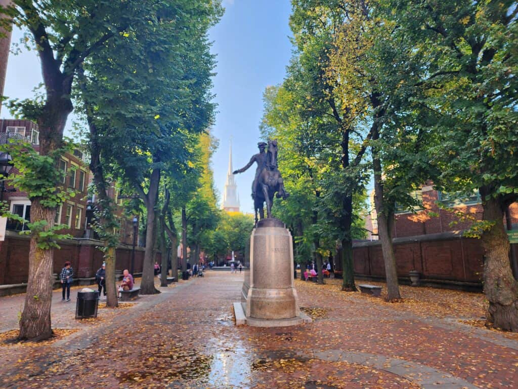 A Boston statue with a row of green trees surrounding and a white steepled church in the distance in Boston, Massachusetts