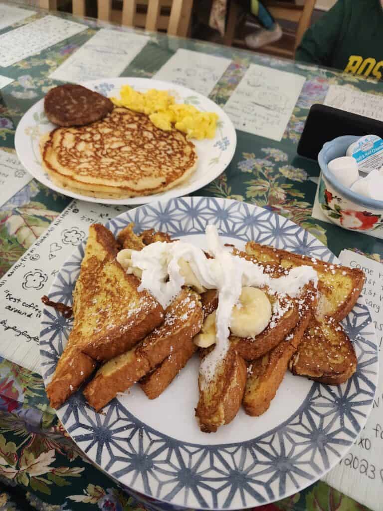 a table with a plate of french toast as well as a plate with a pancake, sausage patty, and scrambled eggs