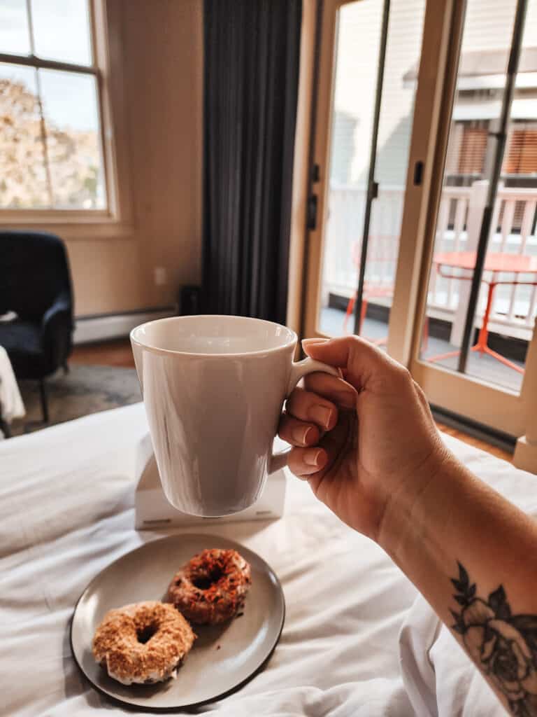 a female arm holds up a white coffee mug with colorful donuts below sitting on a hotel bed