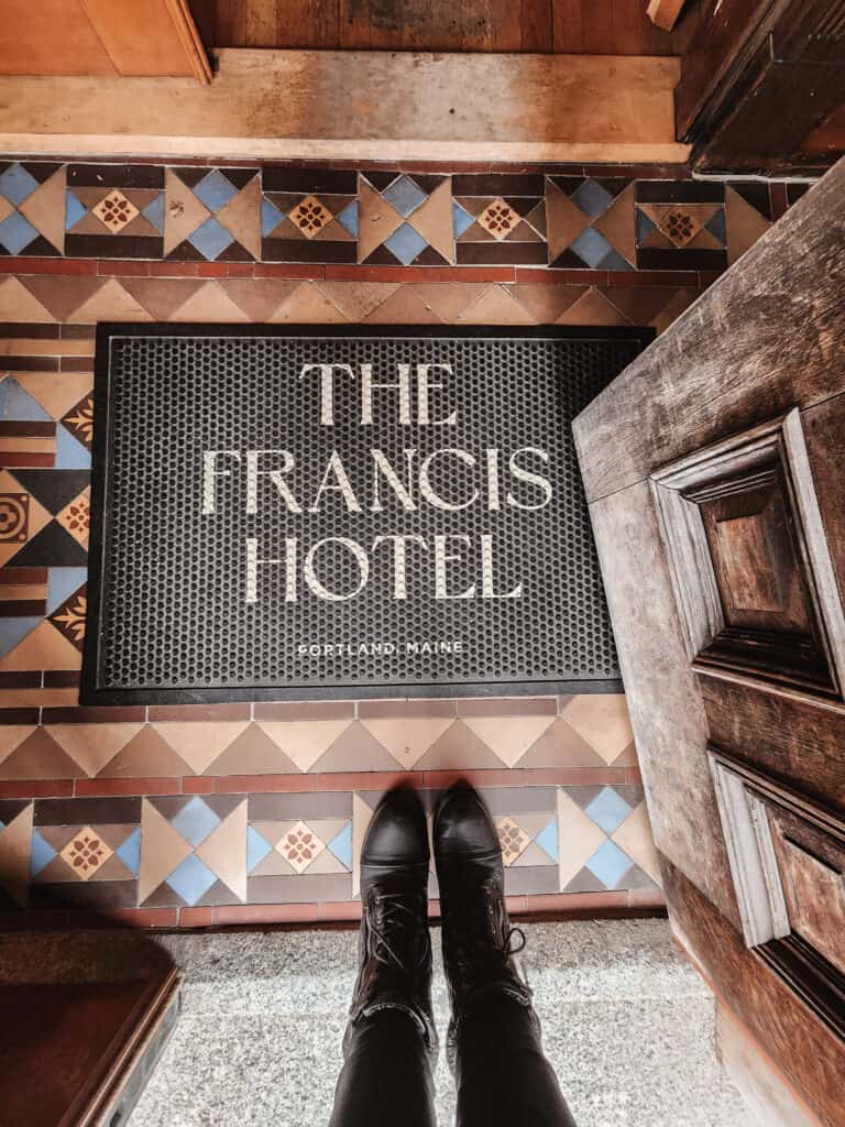 looking down at a welcome mat that reads "the francis hotel portland, maine" with a pair of black boots standing near the bottom, a door is open