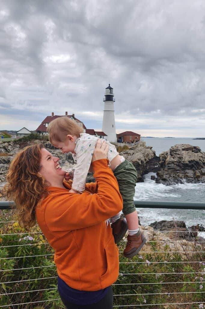 amy is wearing a bright orange sweatshirt and holding her toddler son in the air, both are smiling and there is a famous maine lighthouse beyond