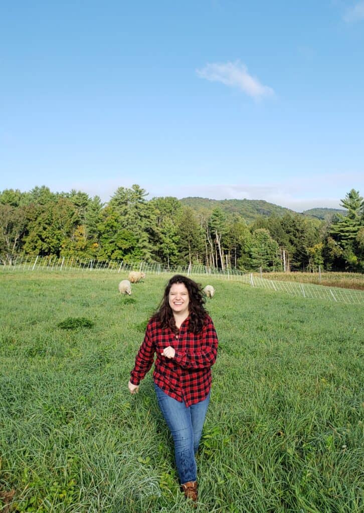 a smiling woman in a red and black plaid flannel frolics in a green field with sheep in the distance - things to do in vermont