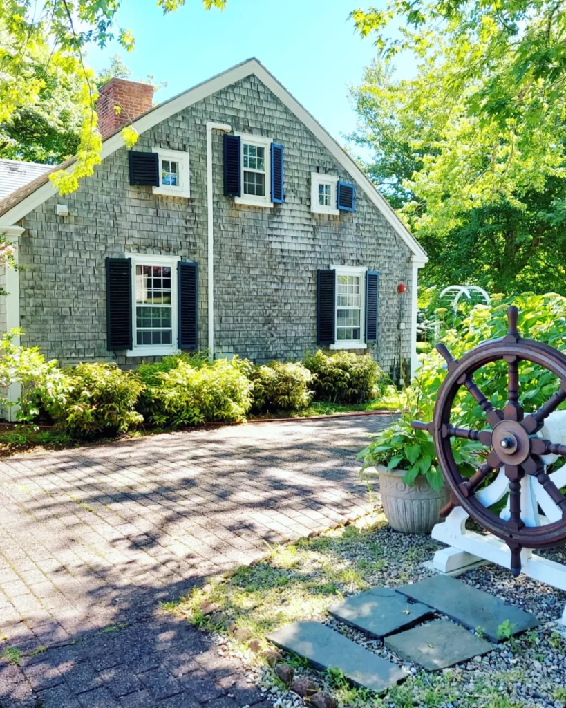 A grey brick Cape Cod structure with a maritime theme wheel in the garden