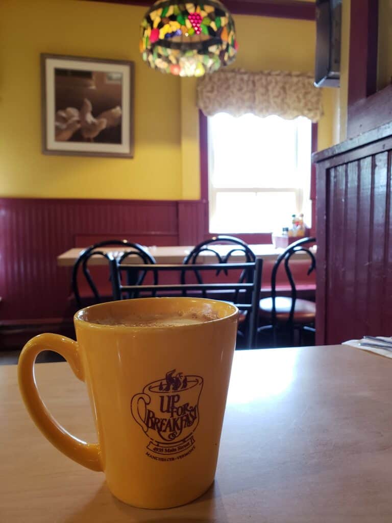 a coffee mug that says "up for breakfast" sits on a table in a cozy cafe with a tiffany lamp chandelier in the back