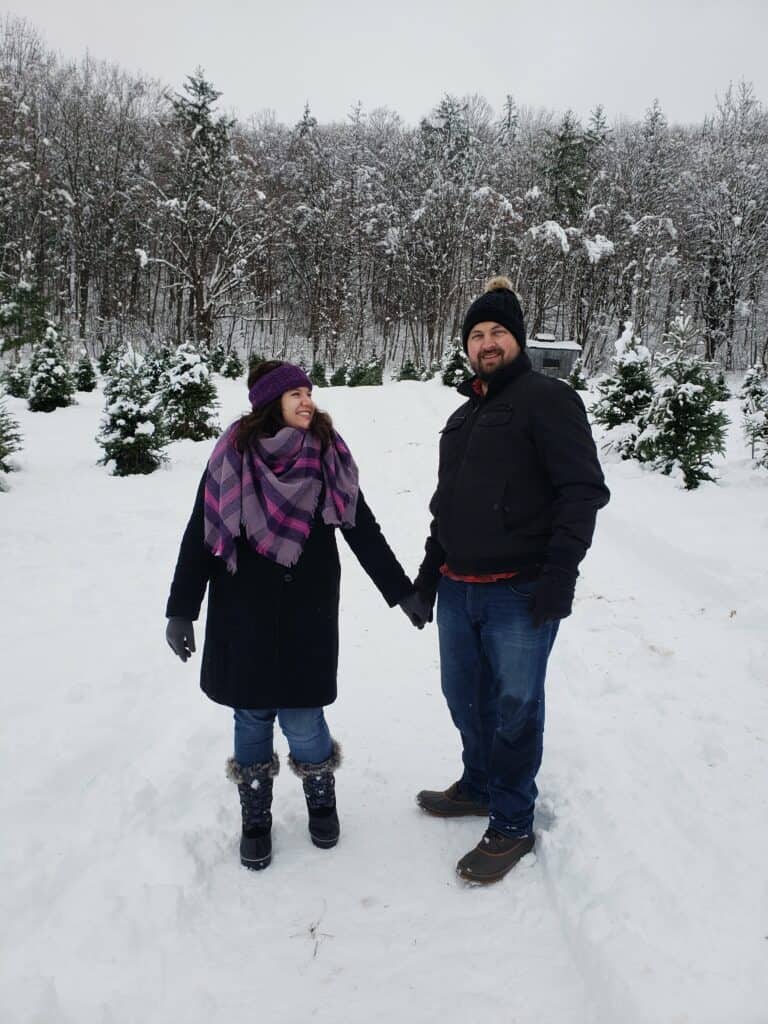 a couple standing in a christmas tree farm holding hands and smiling, it is very snowy