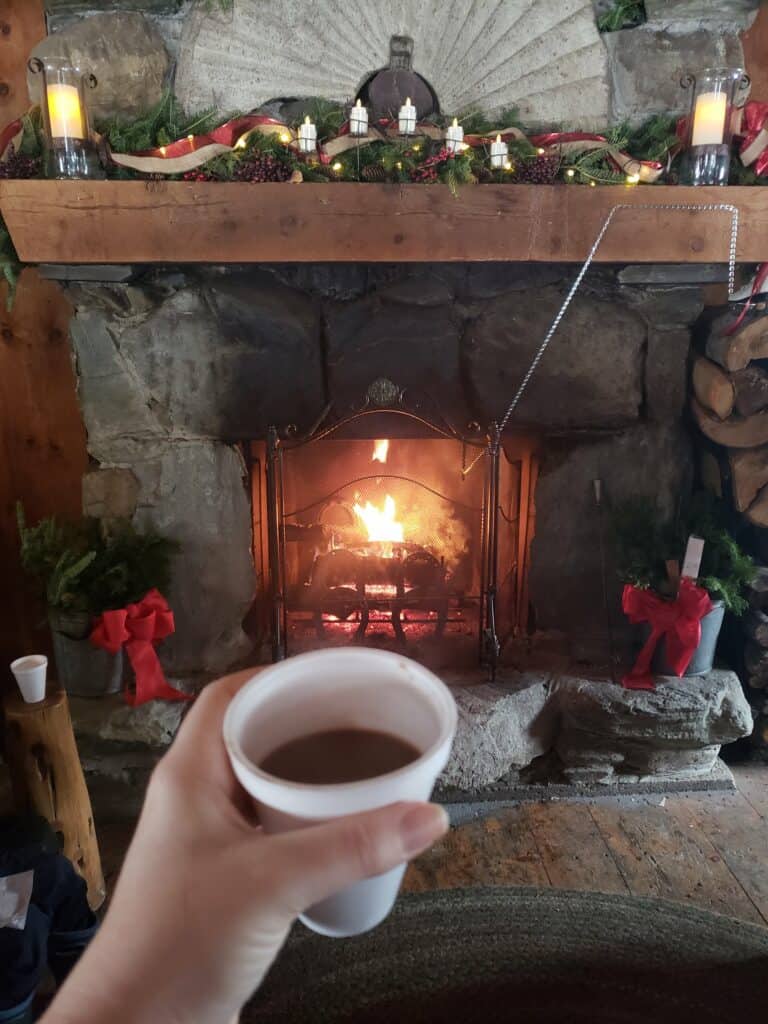 a hand holds a styrofoam cup with hot chocolate in front of a cozy fireplace with christmasdecor