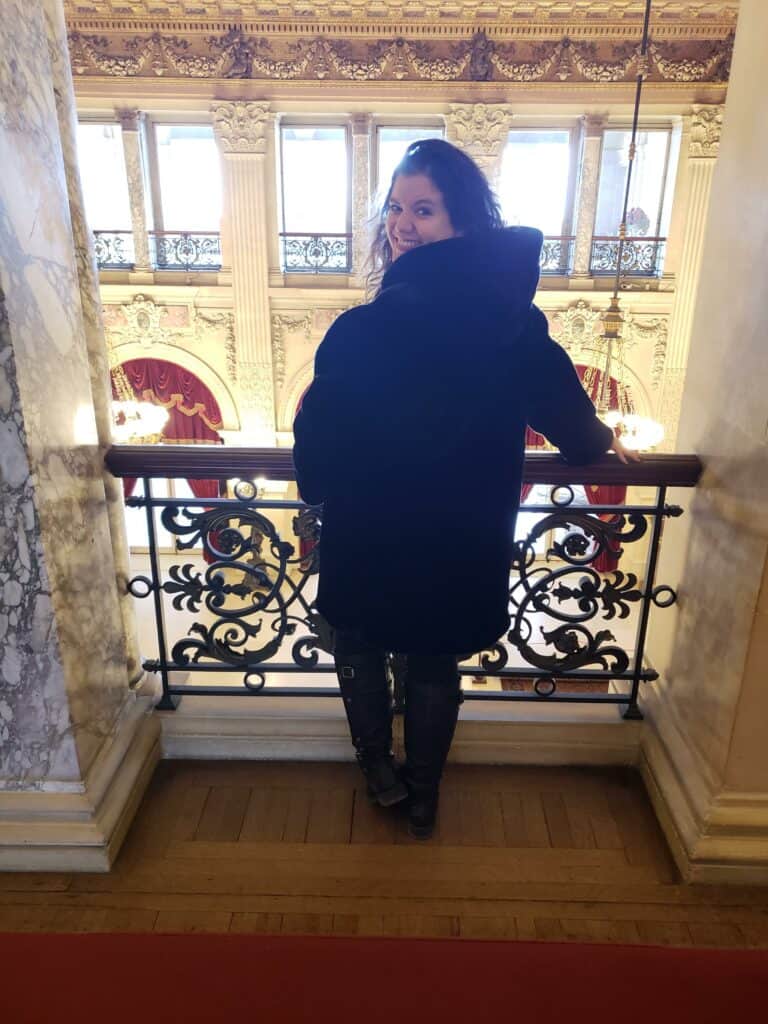 a woman in a black fur coat stands at a balcony overlooking a fancy ballroom and looks over her shoulder and smiles