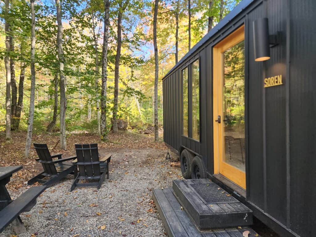 The image showcases a modern, minimalist black cabin named "SOREN" in a forest setting. In front of the cabin, a pair of Adirondack chairs invites guests to relax and enjoy the natural surroundings. The gravel ground and wooden steps to the cabin add to the rustic charm, while the tall trees provide a secluded and intimate atmosphere. 