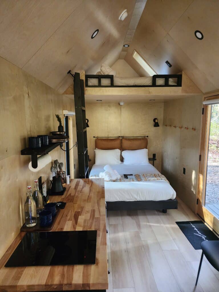 the cozy interior of a minimalist A-frame cabin, with a focus on efficient use of space. The room features a comfortable bed, a lofted sleeping area accessible by a ladder, and a neat kitchenette with modern appliances. The wooden walls and flooring give off a warm, inviting atmosphere, complemented by natural light entering through the large windows.