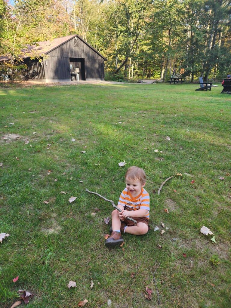 This image captures a moment of childhood joy, featuring a young child sitting on the grass, gleefully playing with a stick. Behind the child, a rustic black barn provides a charming rural backdrop, complete with surrounding trees and a picnic table that hint at leisurely days spent outdoors. The scattered leaves on the lawn suggest the early days of fall, adding a seasonal touch to this wholesome scene