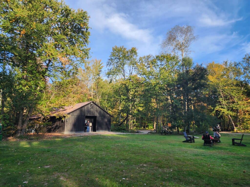  a rustic black barn with an open door, nestled in a sunlit clearing surrounded by dense woodland with early autumn colors. Several wooden Adirondack chairs are arranged on the grass, inviting relaxation in this tranquil outdoor setting. The atmosphere is peaceful, typical of a country retreat or a campground scene
