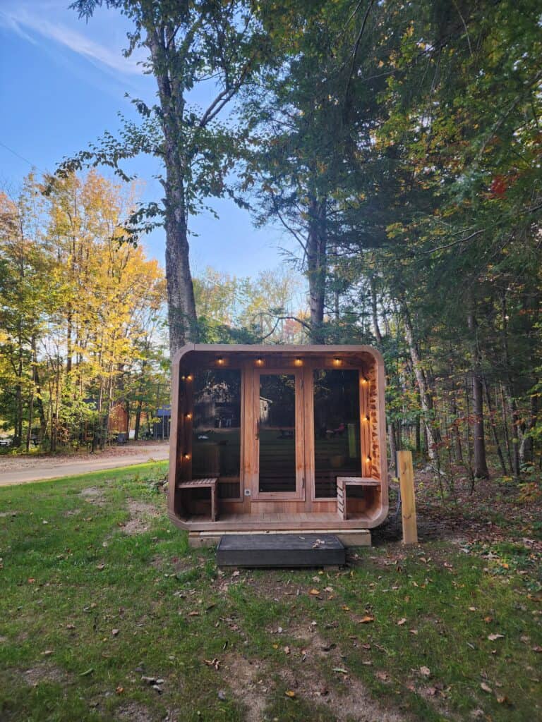 A modern tiny home-style sauna is nestled among trees in early autumn, with leaves changing colors. The sauna features large glass doors, exterior lighting, and a welcoming wooden deck, set against a backdrop of a clear blue sky.