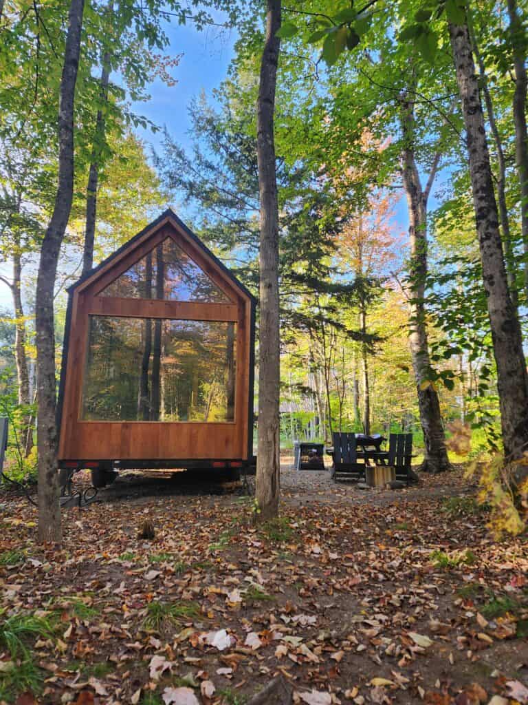 The image presents a charming A-frame micro cabin nestled in a forest setting, with the early signs of autumn visible in the changing leaves scattered on the ground. The cabin's striking glass front provides a reflective surface that captures the surrounding woods, enhancing the sense of seclusion. A pair of dark Adirondack chairs await visitors,