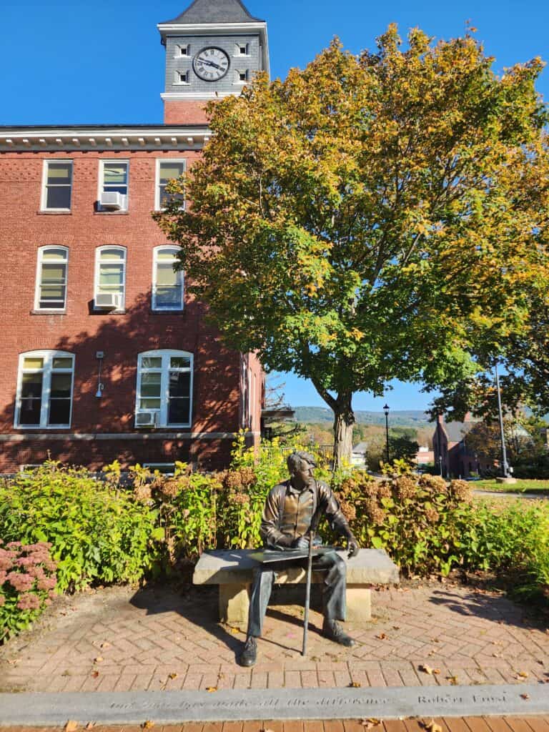 an elegant brick college building rises behind a fall tree and a metal statue of Robert Frost on a bench