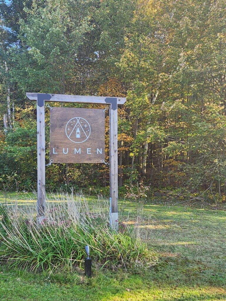 The image features a wooden signboard with the "LUMEN" logo prominently displayed, set against a backdrop of lush greenery. The sign stands at the entrance of what appears to be a nature retreat or park, inviting visitors into the tranquility of the forest beyond. The warm sunlight bathing the scene suggests a welcoming atmosphere for outdoor enthusiasts and travelers.