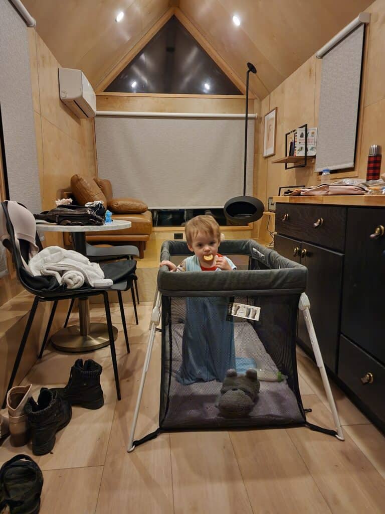 A young child smiles from within a portable playpen in a tidy tiny house living area, surrounded by home comforts like shoes neatly paired by the door, a high chair, and a wooden dresser under warm lighting.