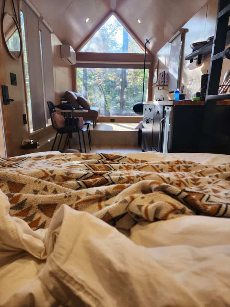 Cozy tiny house cabin interior with a focus on the comfortable bed in the foreground, patterned blanket, and a view of the wooded area through the large window, with a well-equipped kitchenette and sitting area in the softly lit background.
