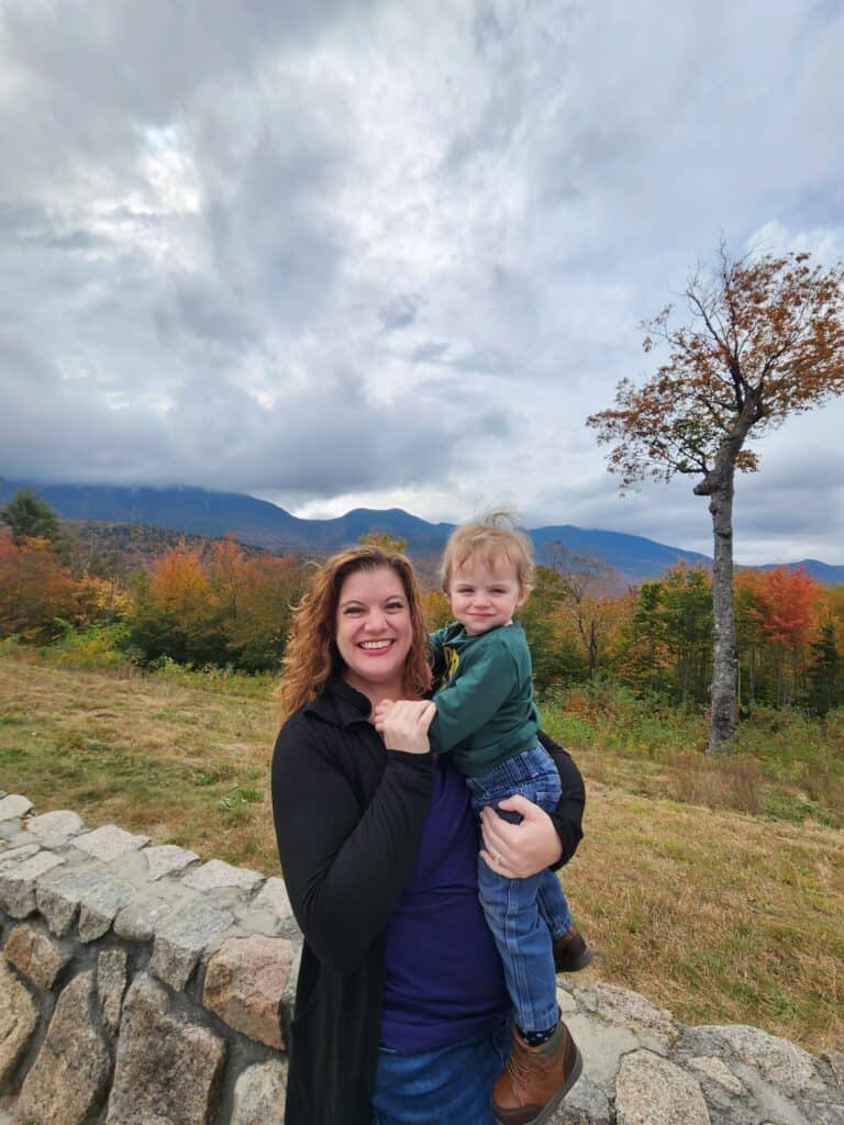 A joyful mother holding her toddler while enjoying a fall trip to New England, with a backdrop of colorful autumn trees and mountainous scenery under a dramatic cloudy sky.