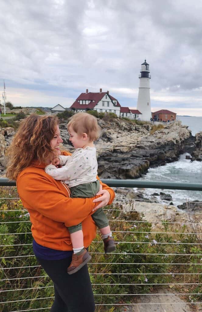 amy, a woman in her 30s, holds her toddler son and they smile at each other with portland head light in the distance, portland maine lighthouse