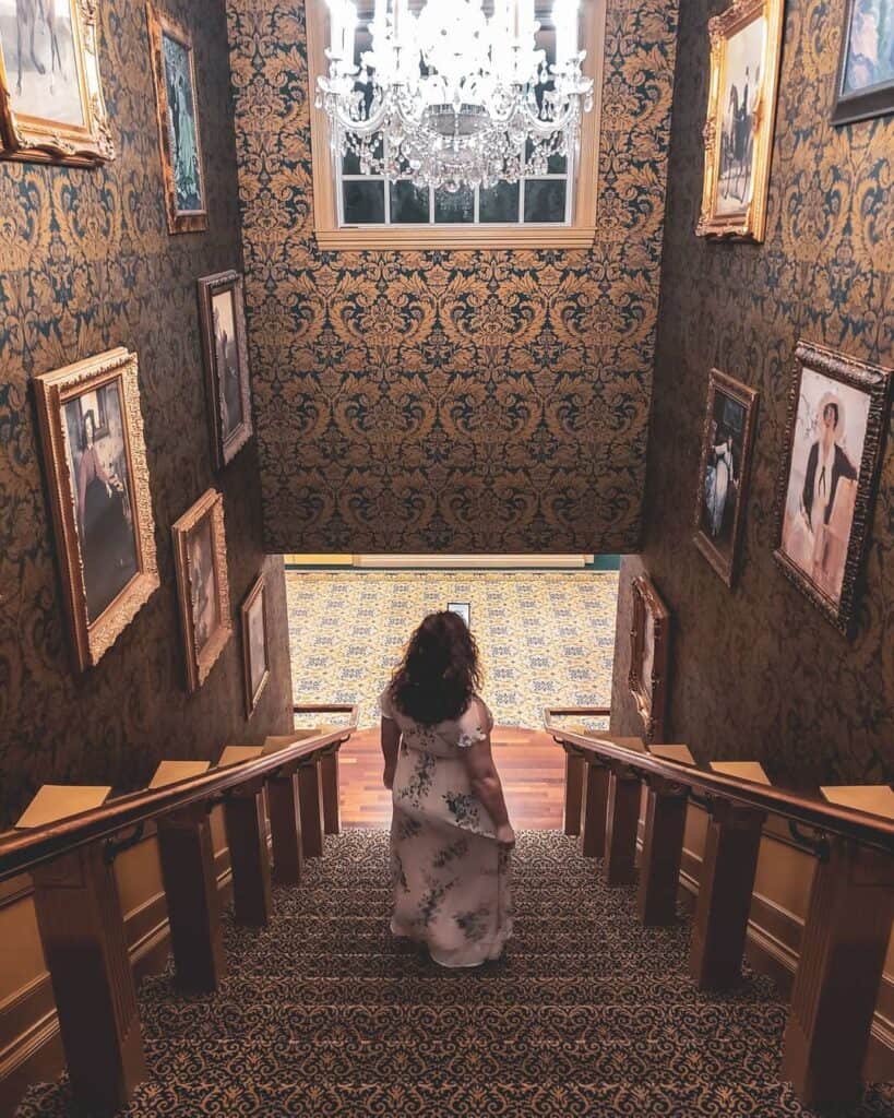 amy walks down the stairs in a hallway with fancy wallpaper and portraits